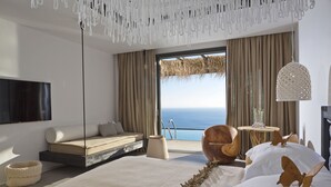 A modern bedroom with a large bed, a sofa, a wooden chair, a hanging light fixture, and a view of the ocean.