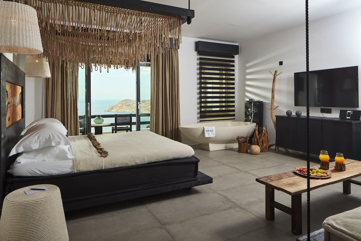 A modern bedroom with a bed, a dining table with breakfast, a TV, and a view of the sea.