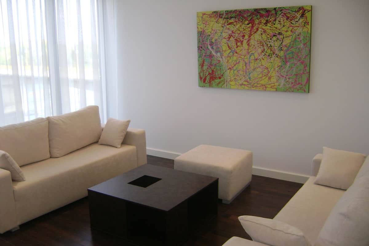 A modern living room with a beige sofa, a matching ottoman, and a dark coffee table. A colorful abstract painting hangs on the wall.
