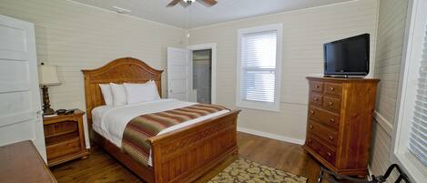 Un dormitorio con cama de madera, mesita de noche, cómoda, televisor encima y ventilador de techo, además de una ventana con persianas.