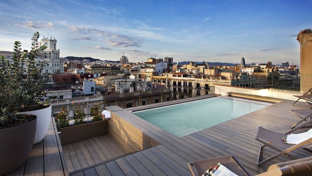 A rooftop pool with wooden deck and lounge chairs, overlooking a cityscape with historic buildings. - Hotel Vincci Gala (Barcelona)