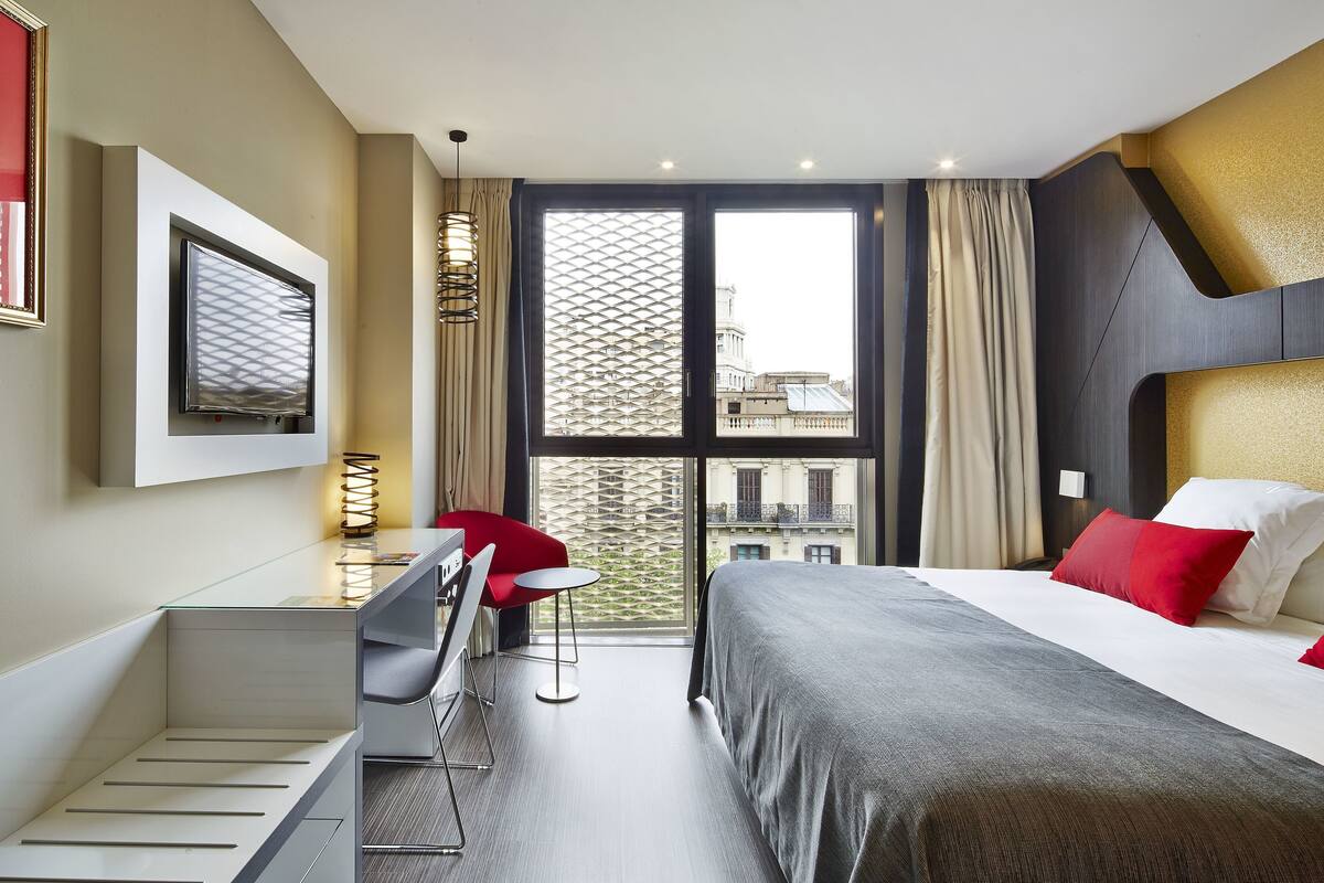A modern hotel room with a bed, a desk, a chair, and a large window with a view of buildings.