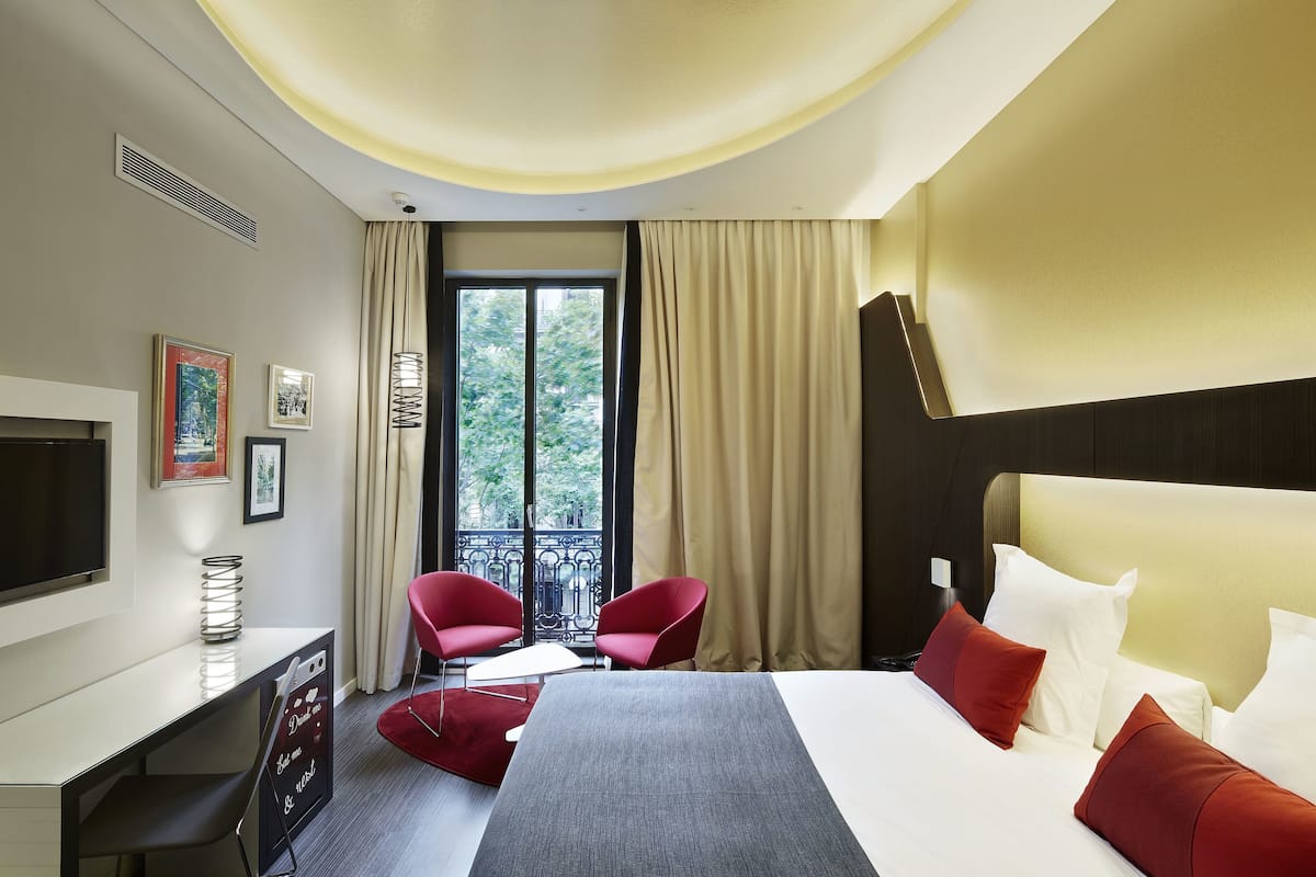 A hotel room with a bed, a desk, a TV, red chairs, a balcony with curtains, and a view of trees.