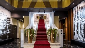A grand staircase with a red carpet, flanked by large potted plants, leading to a high-ceilinged area with patterned walls and a large mural. - Hotel Vincci Gala (Barcelona)
