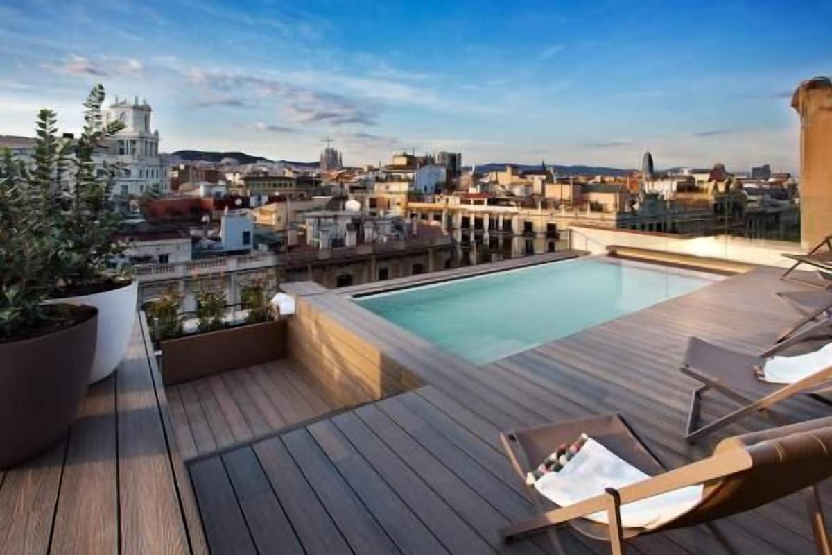 A rooftop pool area with wooden decking, lounge chairs, and a view of the cityscape.