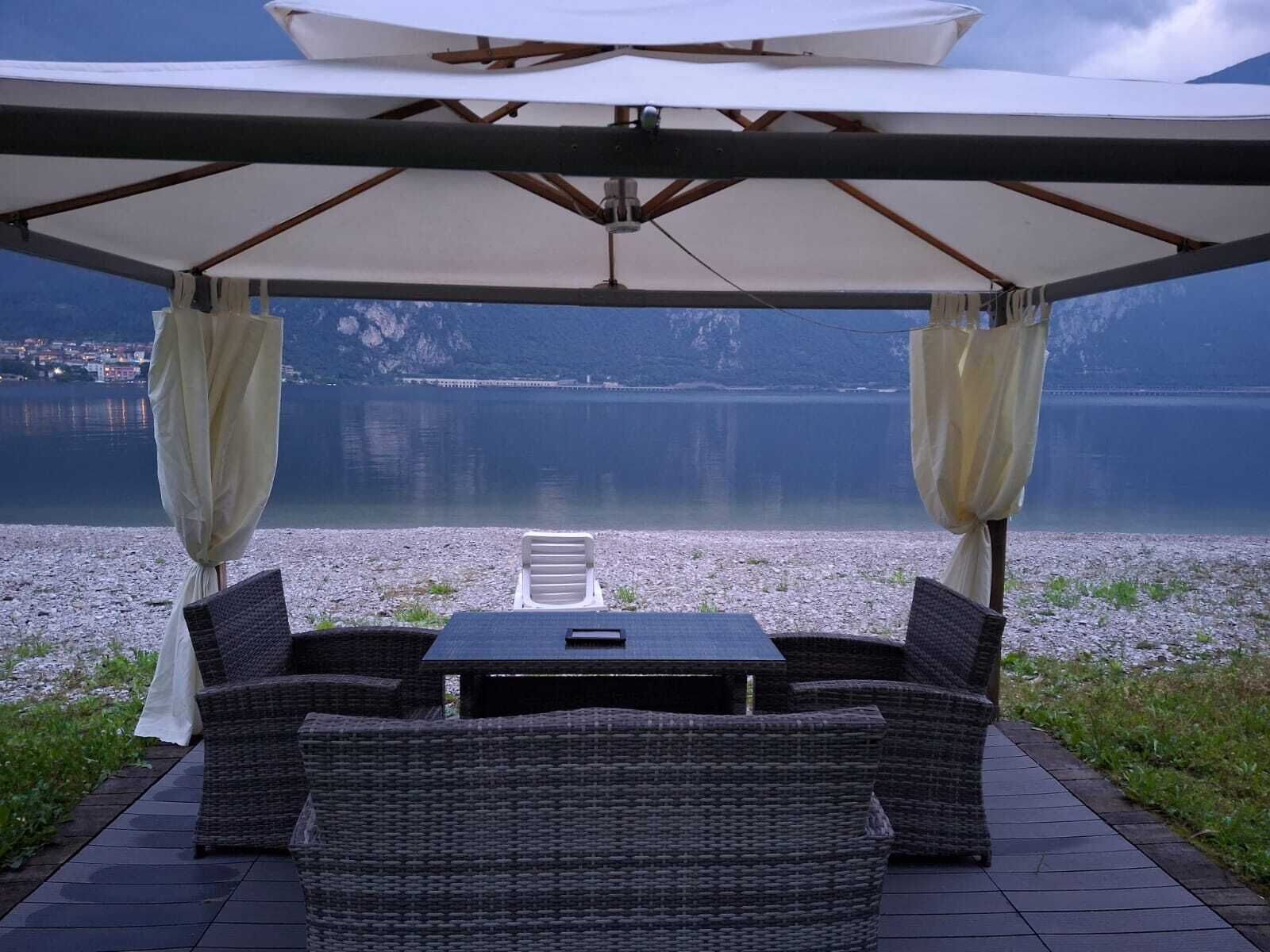 Zona relax all'aperto con mobili in vimini e un tavolo sotto un grande ombrellone, con vista su un lago tranquillo e montagne lontane.
