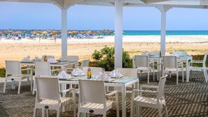 An outdoor dining area with tables and chairs, overlooking a beach with umbrellas and lounge chairs.