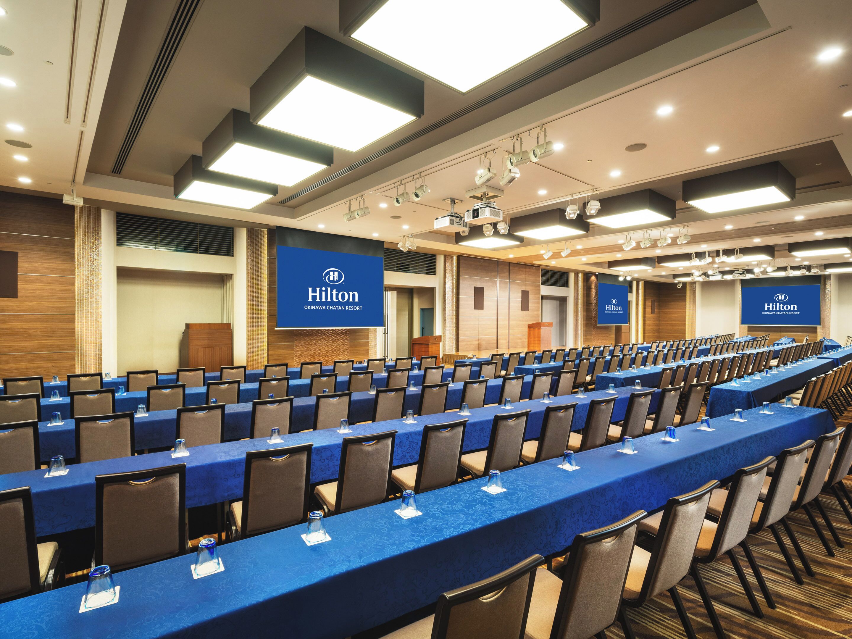 A large conference room with rows of tables and chairs, a podium, and a large screen displaying the Hilton logo.