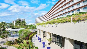 A modern hotel with a curved facade, outdoor seating area with blue tables, and a landscaped garden.
