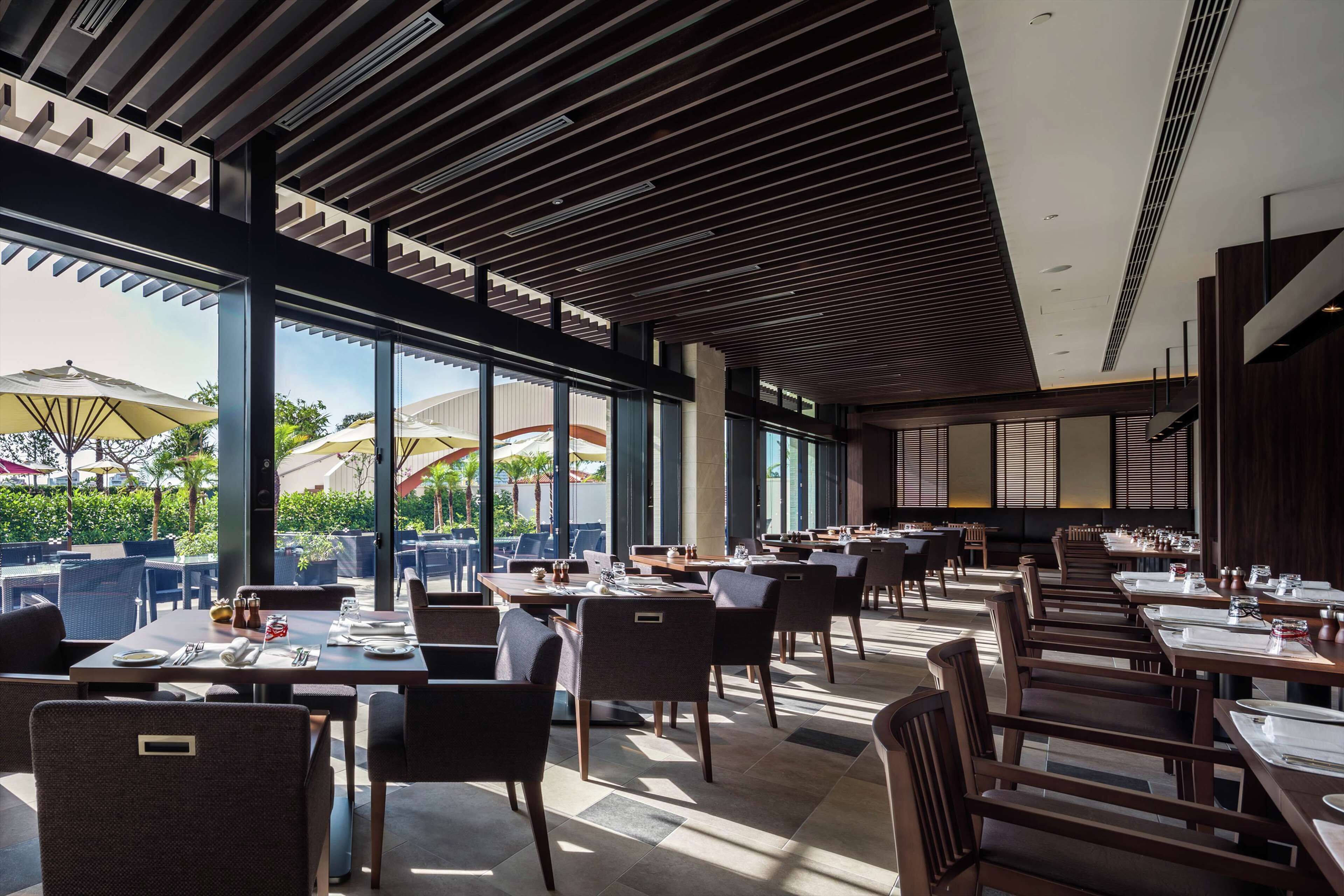 A modern restaurant with a high ceiling, large windows, and outdoor seating visible through the glass.