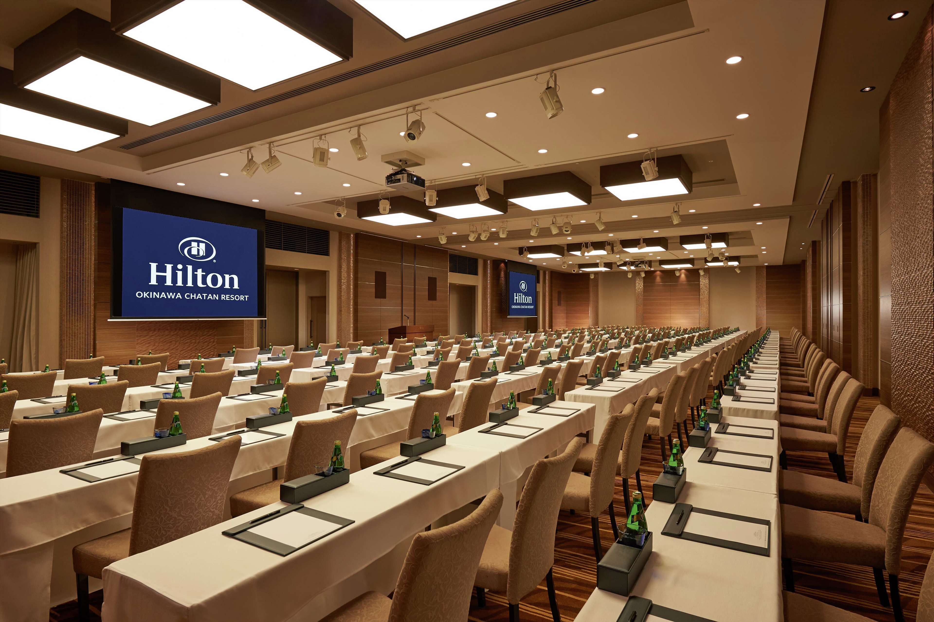 A conference room at the Hilton Okinawa Chatan Resort with rows of tables, chairs, and a large screen displaying the hotel's name.