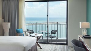 Quarto de hotel com cama, escrivaninha, cadeira, mesa e vista para o oceano por uma porta de correr.