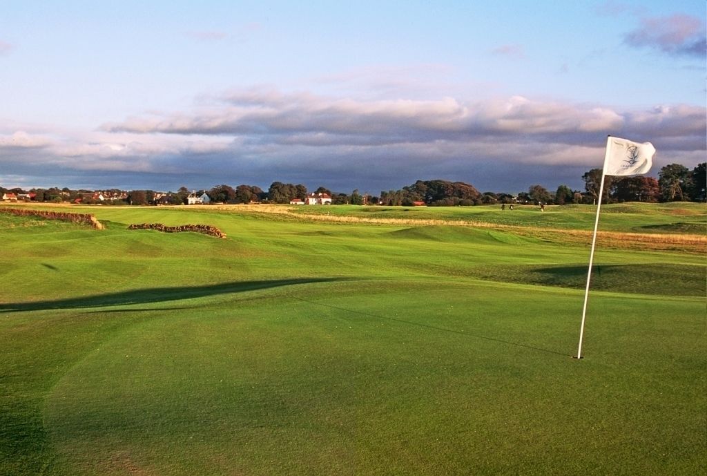Een golfbaan met een vlag op een tee, omgeven door huizen en bomen onder een bewolkte lucht.
