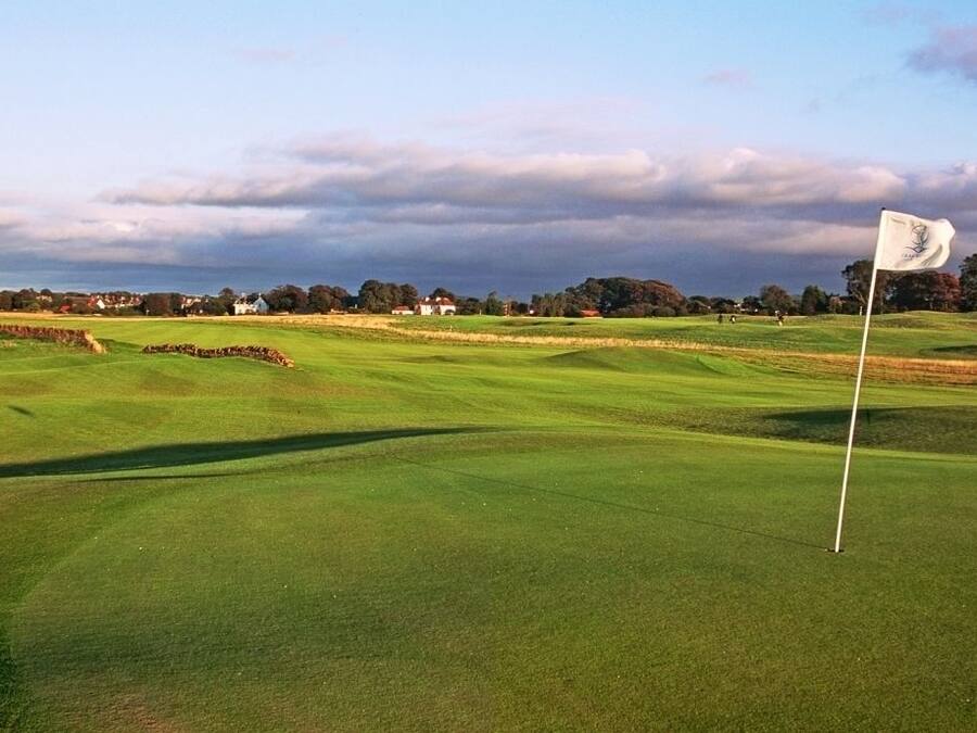 Campo de golf con una bandera en el tee, rodeado de casas y árboles bajo un cielo nublado.