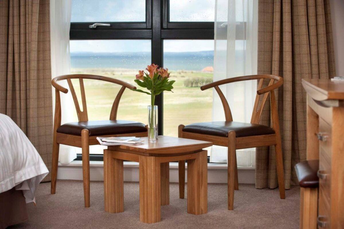Una habitación con dos sillas de madera, una mesita y un jarrón con flores. Se ve una ventana con vistas al exterior.