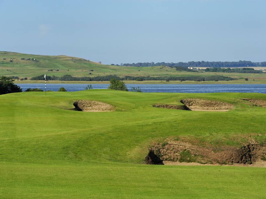 A golf course with bunkers, a flag, and a water body in the background.