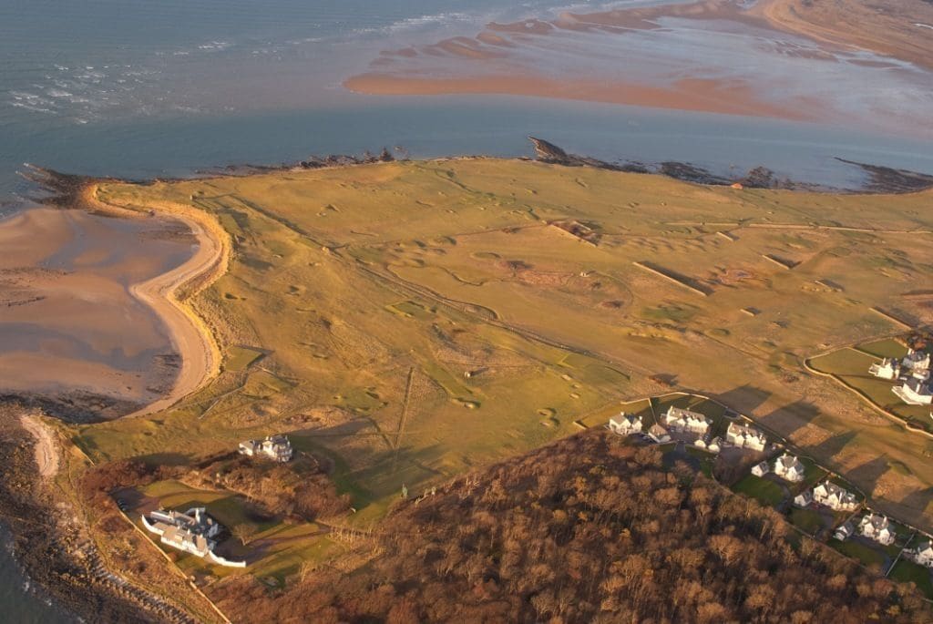 Une vue aérienne d’un paysage côtier, avec une série de bâtiments blancs, probablement un complexe hôtelier ou un hôtel, situé sur une colline surplombant la mer.