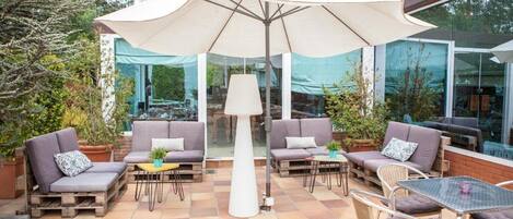 An outdoor patio with cushioned seating, a central umbrella, and a glass-enclosed area in the background.
