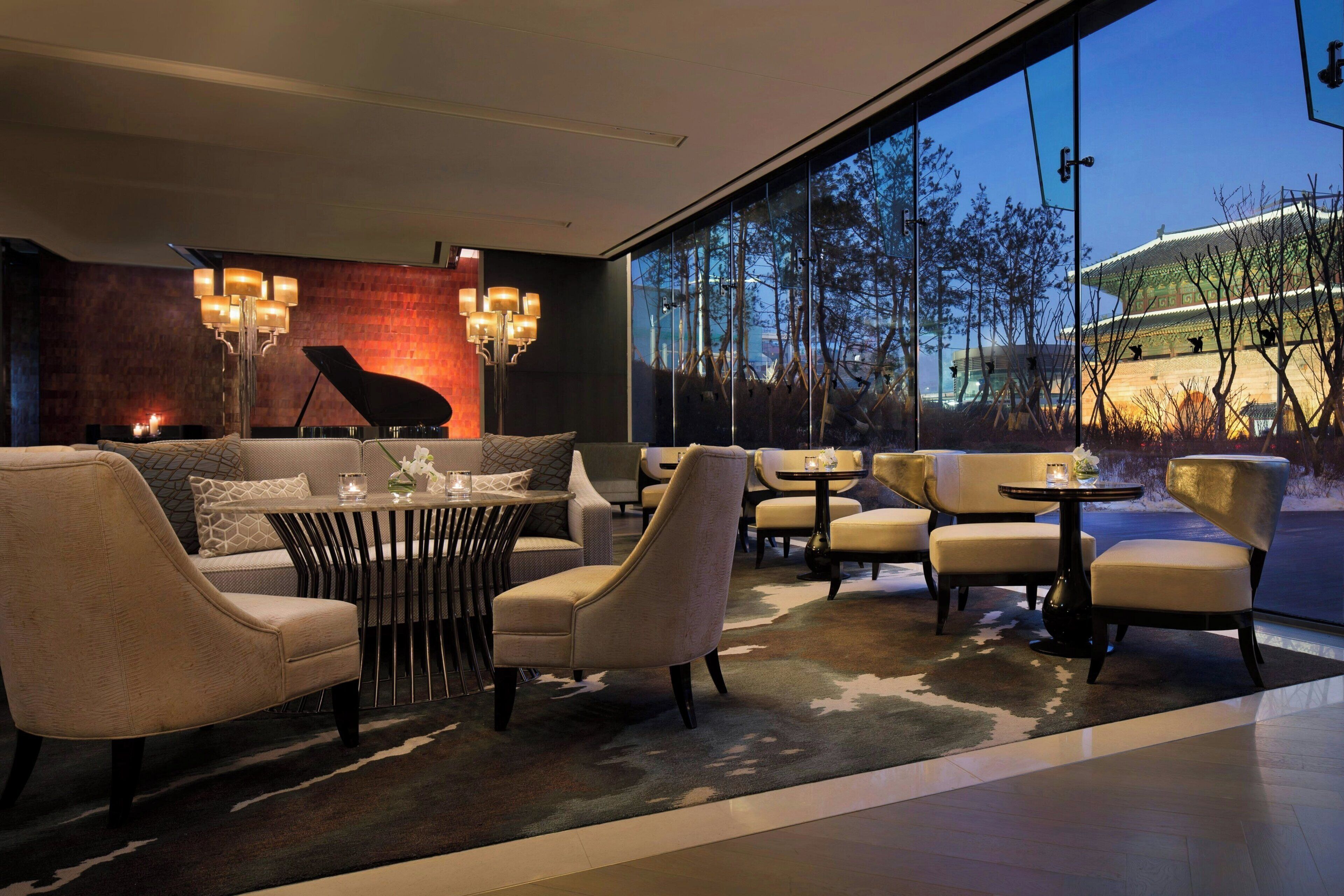 A modern interior with a piano, sofas, and tables, large windows with a view of a traditional building.