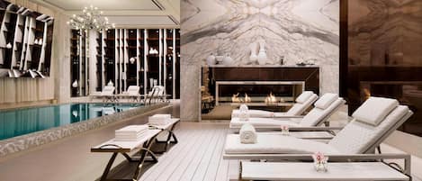 Un área de piscina interior lujosa, con sillones reclinables, chimenea y pared de mármol.