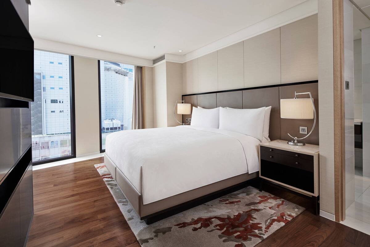 Une chambre d’hôtel moderne dotée d’un grand lit, d’un téléviseur à écran plat, d’une table de chevet avec des lampes et offrant une vue sur la ville grâce à de grandes fenêtres.