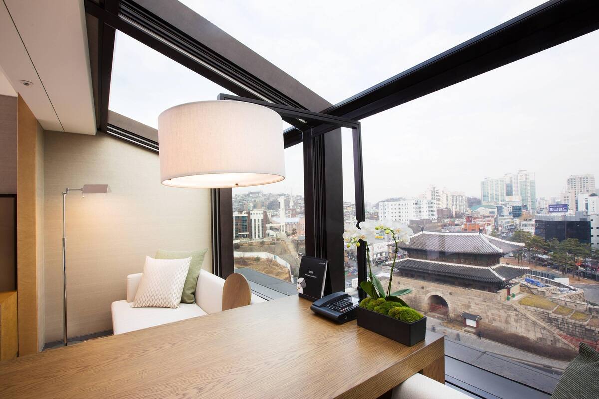 Une chambre d’hôtel moderne dotée d’une grande fenêtre donnant sur la ville, d’un bureau en bois, d’une lampe et d’une petite plante en pot.