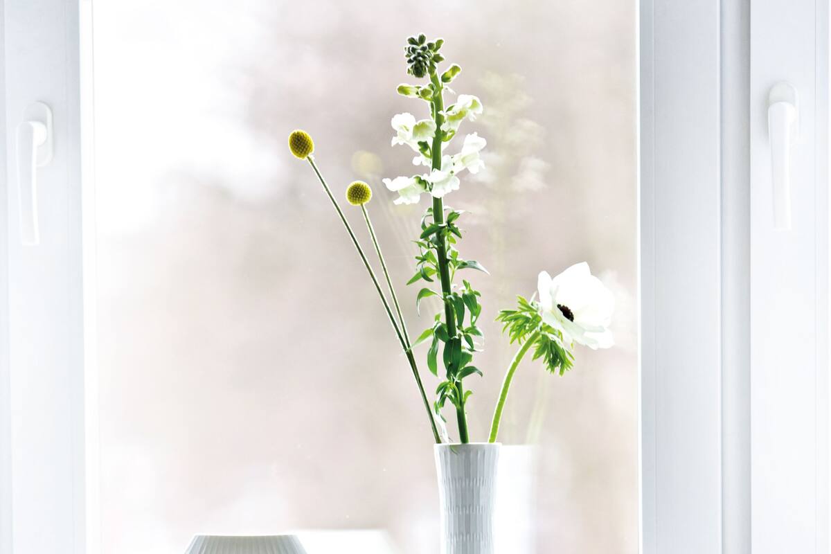 A white radiator, a white vase with flowers, and a white chair by a window.