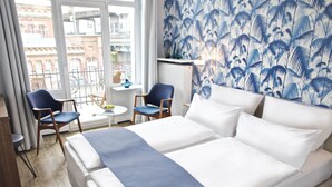 Quarto de hotel com uma cama grande, duas cadeiras, uma mesinha e vista da cidade por grandes janelas.