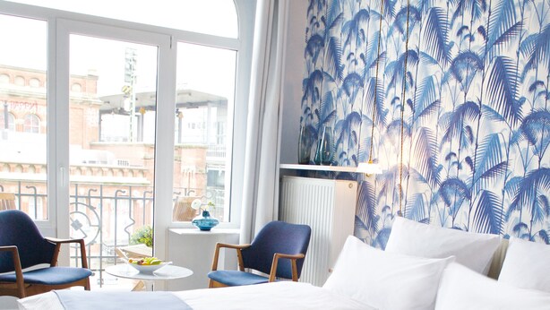 Quarto de hotel com uma cama grande, duas cadeiras, uma mesinha e vista da cidade através de amplas janelas.