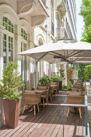 An outdoor seating area with wicker chairs and a large umbrella, surrounded by potted plants and a wooden deck. - PortoBay Liberdade (Lisbon)