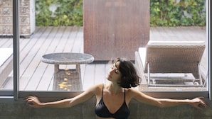 Una mujer con traje de baño negro en una piscina con vistas a un jardín y mobiliario exterior.