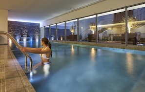 A modern indoor swimming pool with a glass barrier, a woman in a swimsuit, and a view of a building with a staircase and plants. - PortoBay Liberdade (Lisbon)