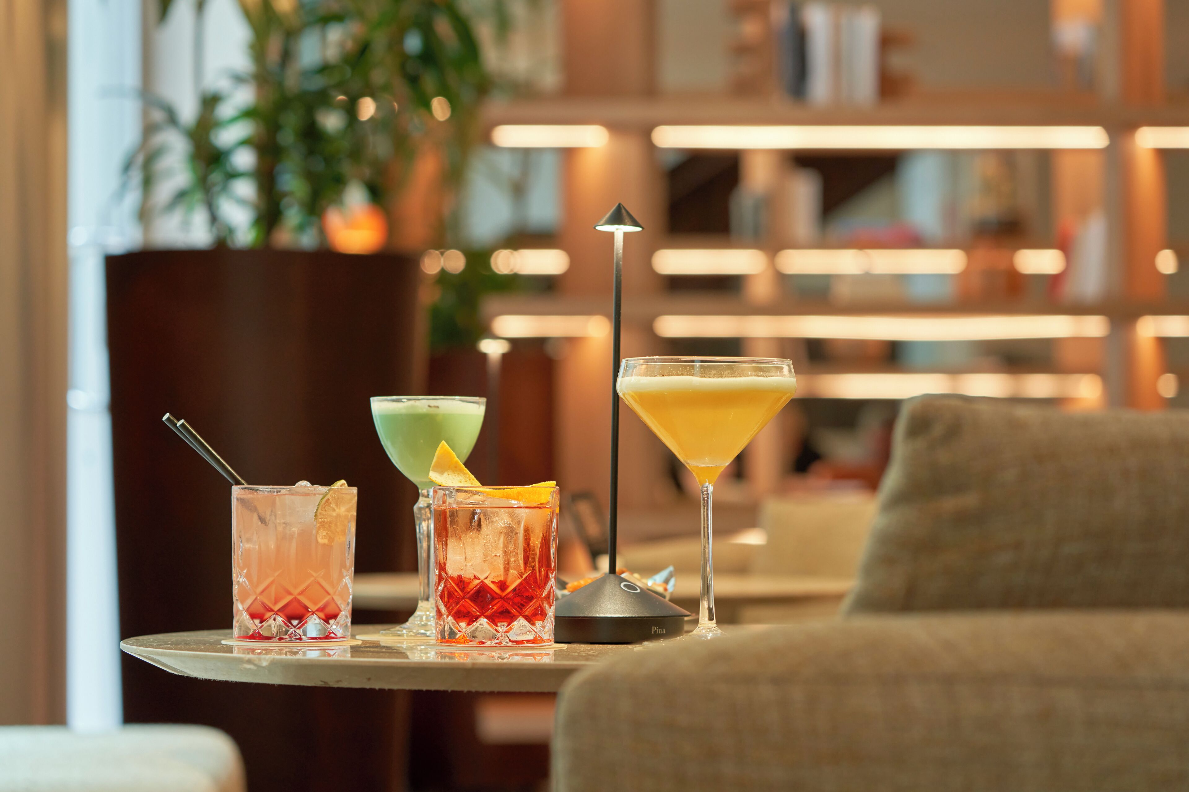 Zona de bar con tres cócteles sobre una mesa, un taburete y una estantería al fondo.
