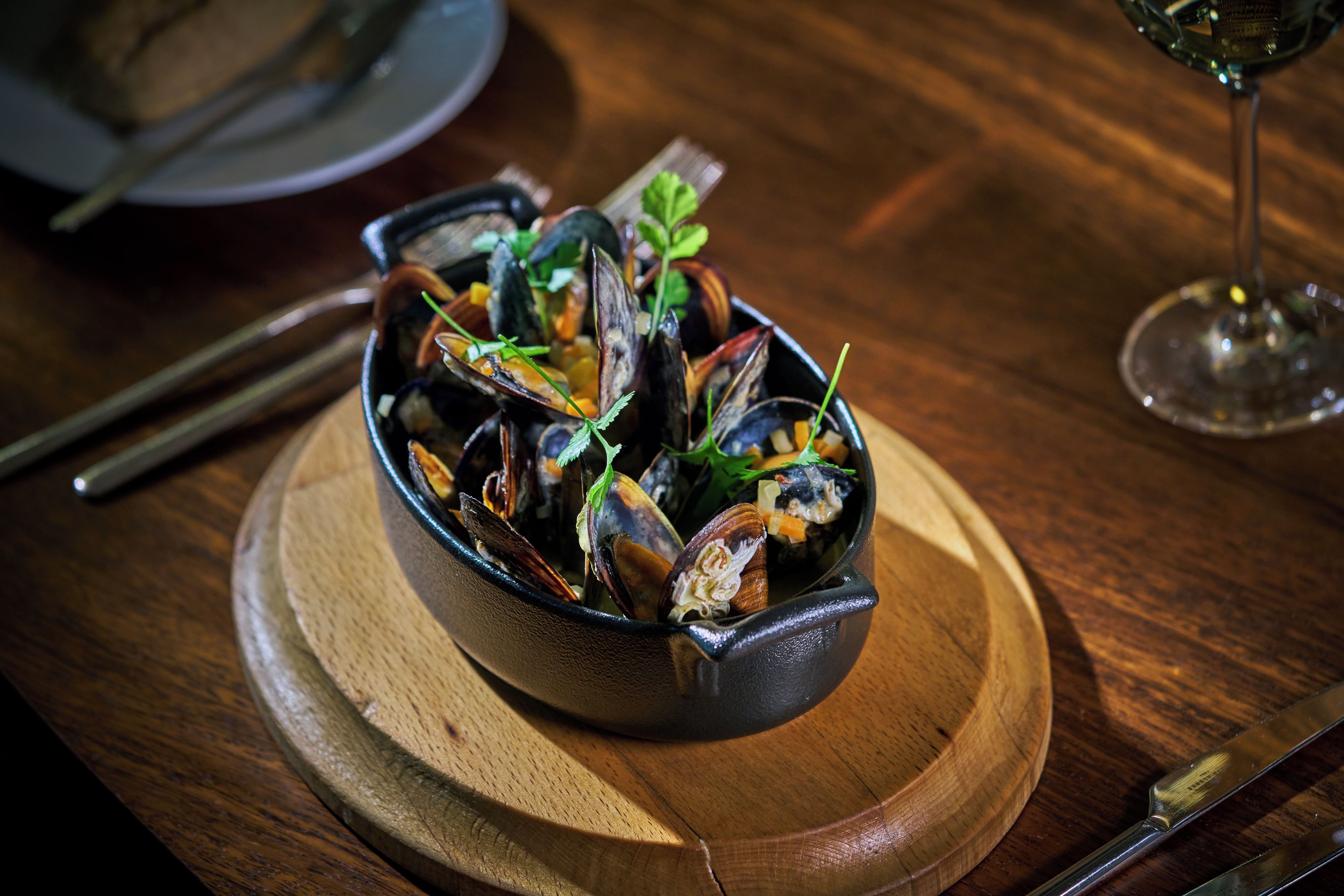 Un bol de moules sur une table en bois, avec un verre à vin et des couverts.