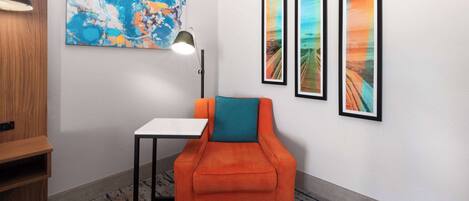 A room with a teal cushioned armchair, a small white table, a lamp, and three framed pictures on the wall.