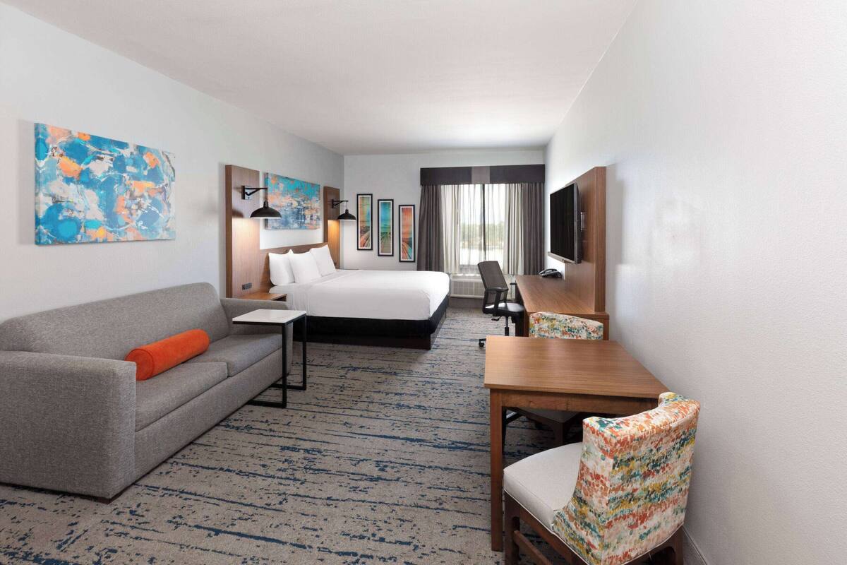 Une chambre d’hôtel avec un lit, un canapé gris orné d’un coussin orange, une table en bois, une chaise à motifs et un téléviseur.