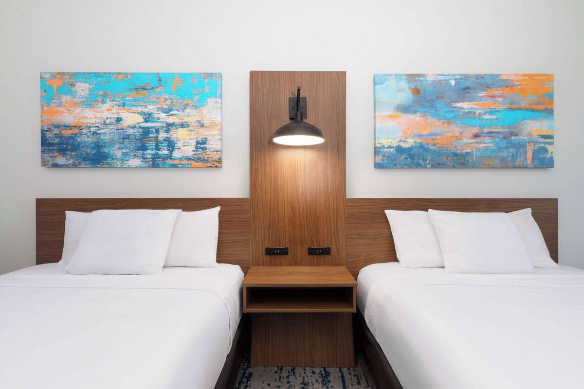 Une chambre d’hôtel avec deux lits, une tête de lit en bois, une lampe fixée au mur et des tableaux abstraits accrochés aux murs.