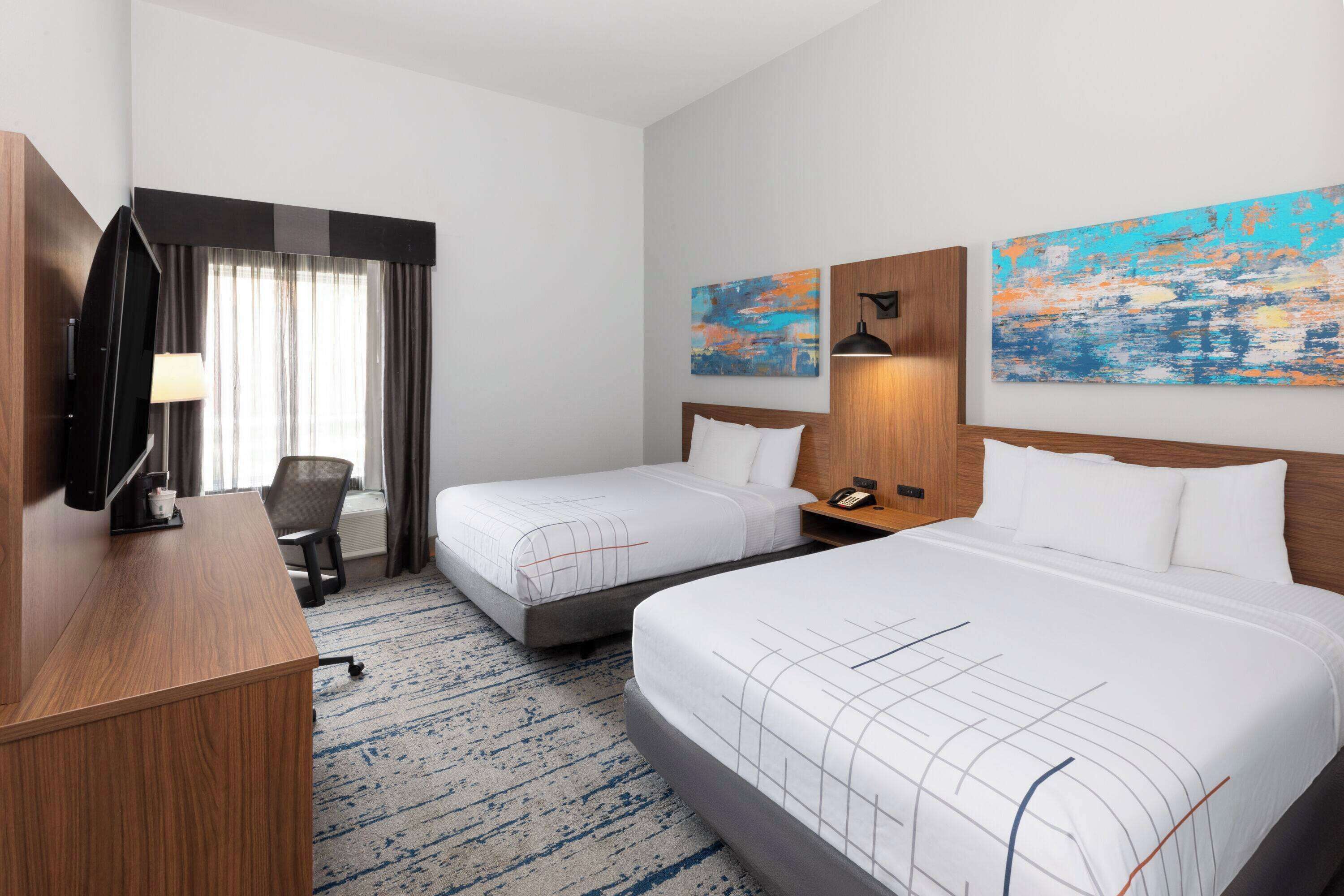A hotel room with two beds, a desk, a TV, and artwork on the walls.