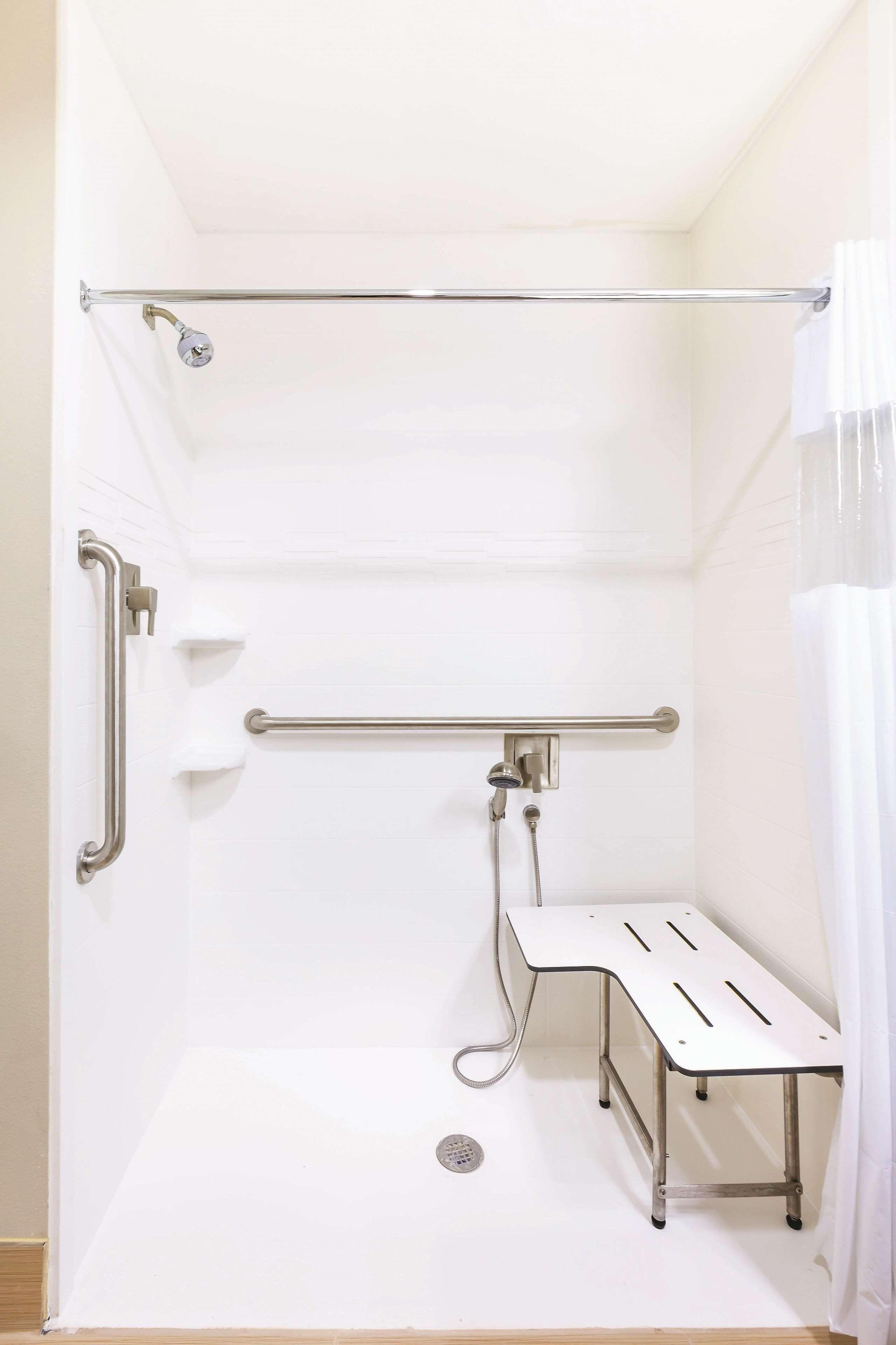 A white, enclosed shower area with a grab bar, a shower seat, and a handheld showerhead.