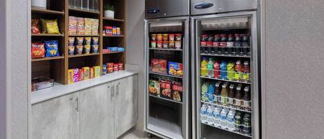 A kitchenette with a refrigerator stocked with beverages and snacks, and shelves with various food items.