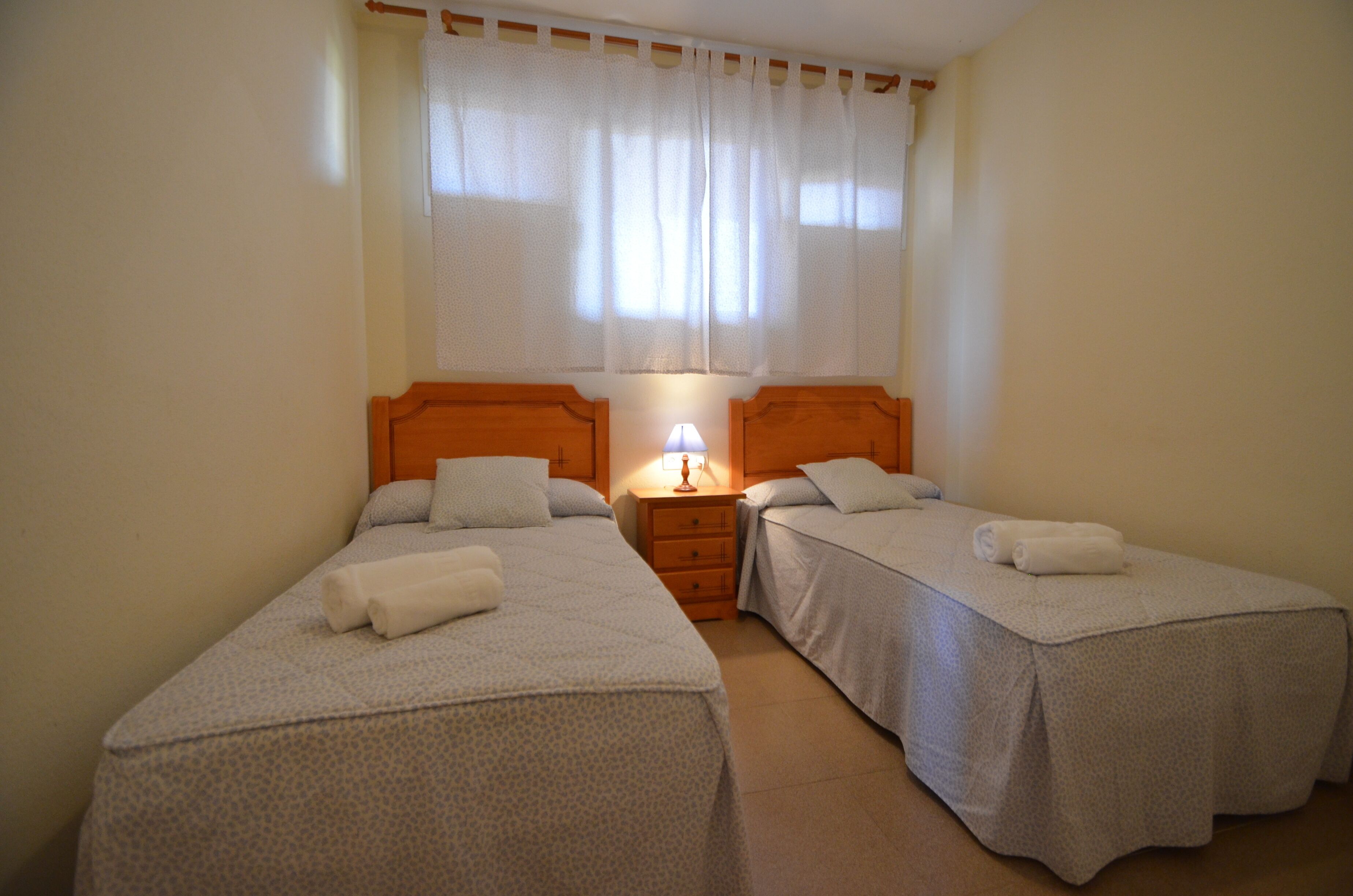 A twin room with two beds, a nightstand with a lamp, and a window with curtains.