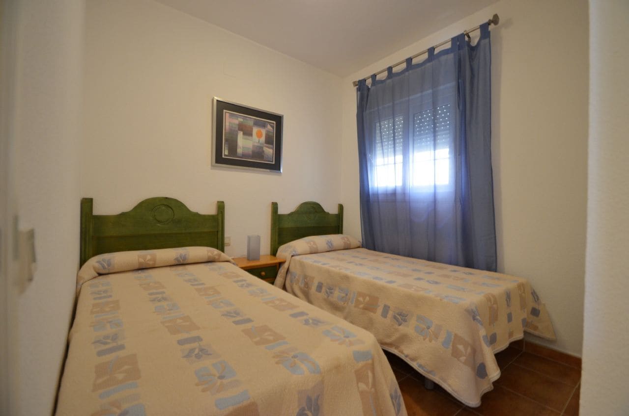 Ein Zimmer mit zwei Einzelbetten, einem Nachttisch, einem gerahmten Bild an der Wand und einem Fenster mit blauen Vorhängen.