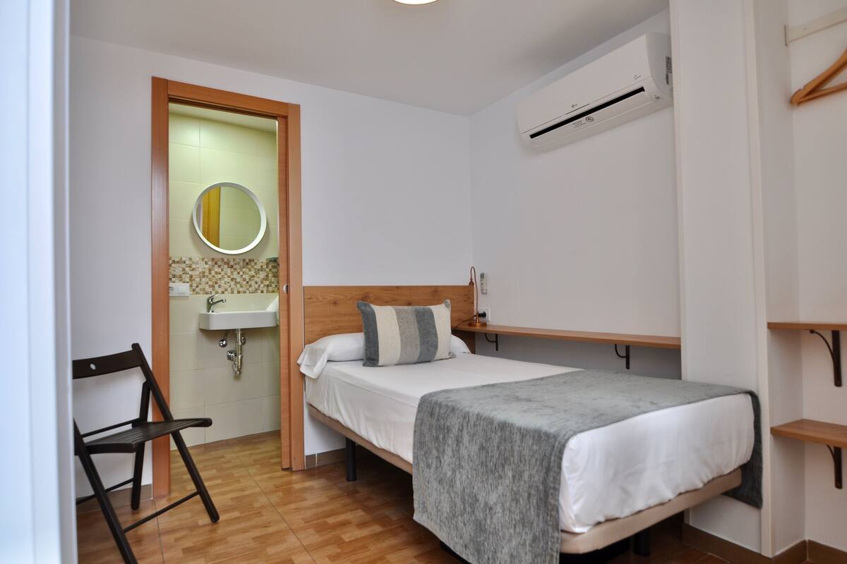 Ein kleines Schlafzimmer mit einem Einzelbett, einem Holzstuhl, einem kleinen Waschbecken und einer Klimaanlage.