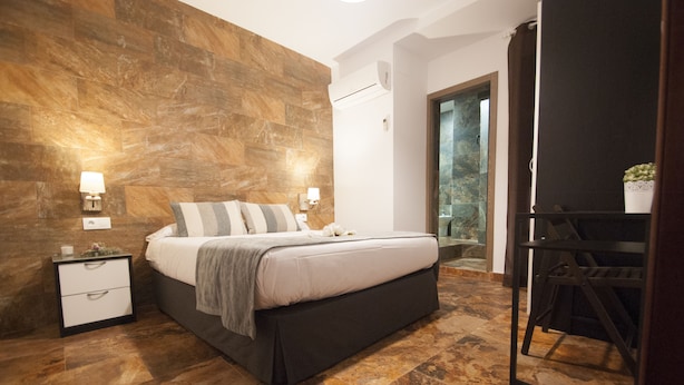 Ein modernes Schlafzimmer mit Steinwand, einem Bett mit grau-weiß gestreiftem Kopfkissen, einem Nachttisch und einem kleinen Tisch mit einer Pflanze.