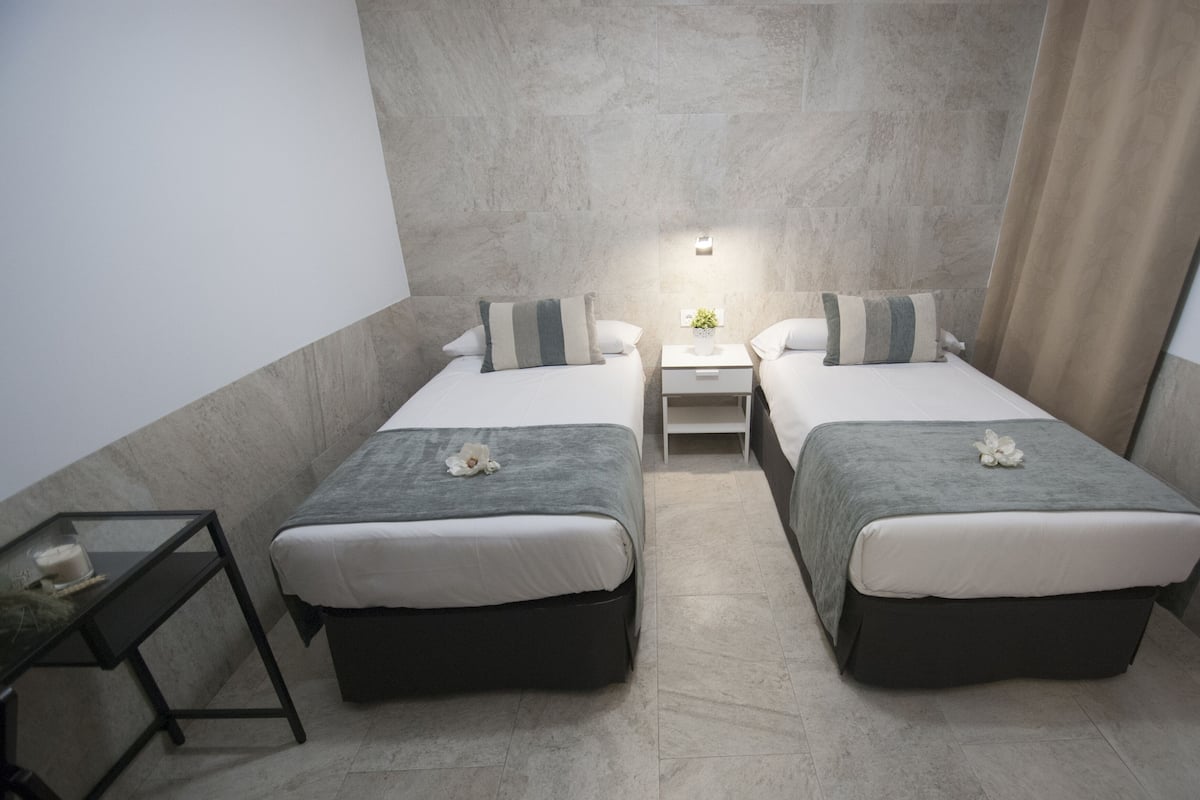 Ein modernes Hotelzimmer mit zwei Einzelbetten, einem kleinen Nachttisch mit Lampe und einem Beistelltisch mit Pflanze.