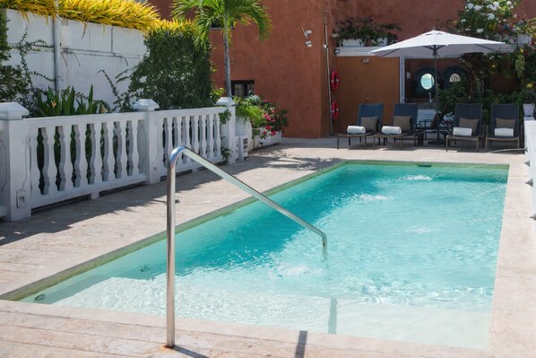 A rectangular swimming pool with a metal ladder, surrounded by a white fence, with lounge chairs and umbrellas in the background.
