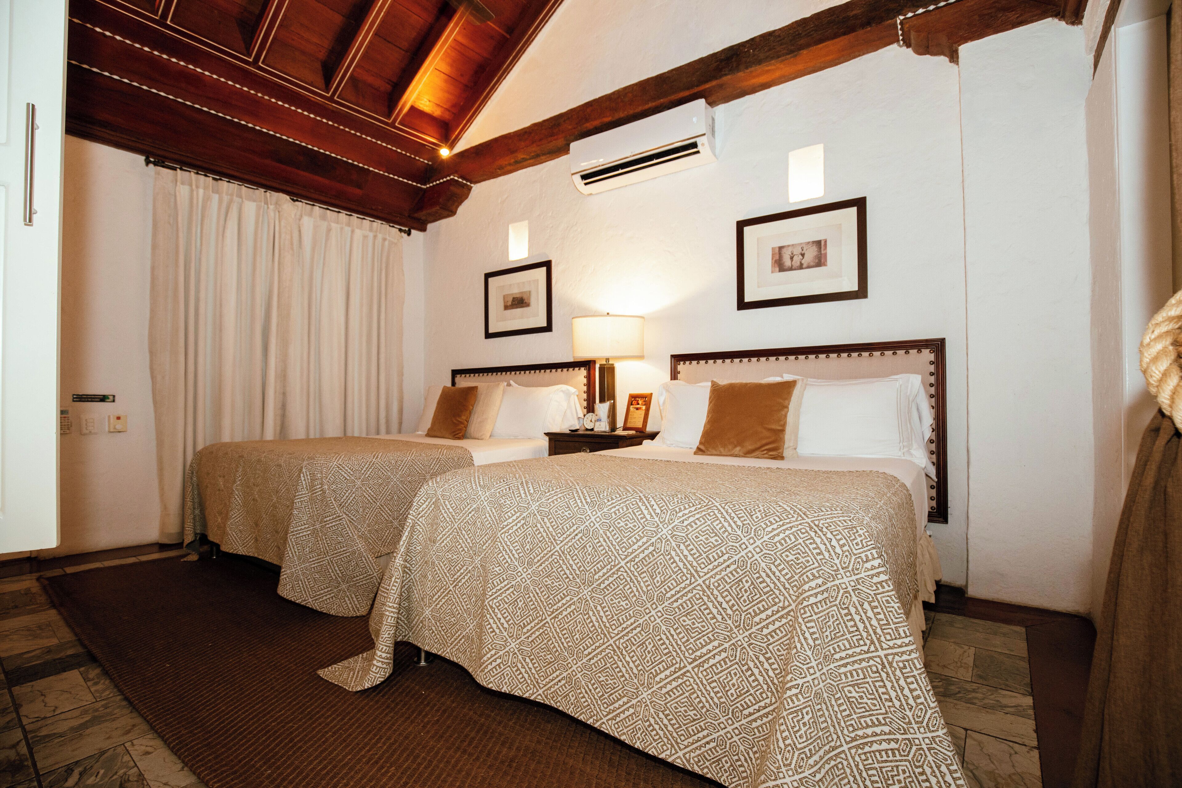 A hotel room with two beds, a wooden ceiling, an air conditioner, and framed pictures on the wall.