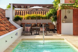 Ein Poolbereich mit einer gefliesten Dachkonstruktion, Außenmöbeln und einer Glocke, die an einer Holzstruktur hängt.
