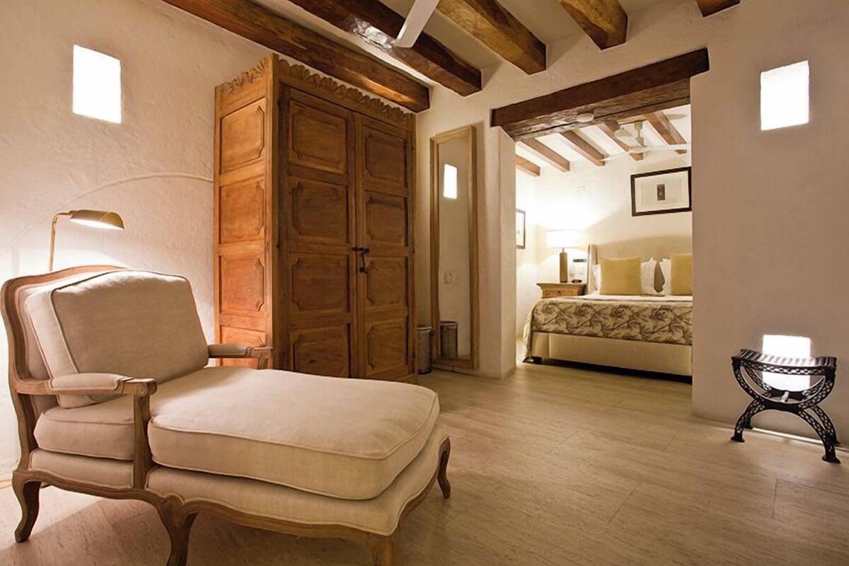 Une pièce comprenant une grande armoire en bois, un fauteuil rembourré, une petite table et un lit recouvert d’une couvre-lit à motifs floraux.
