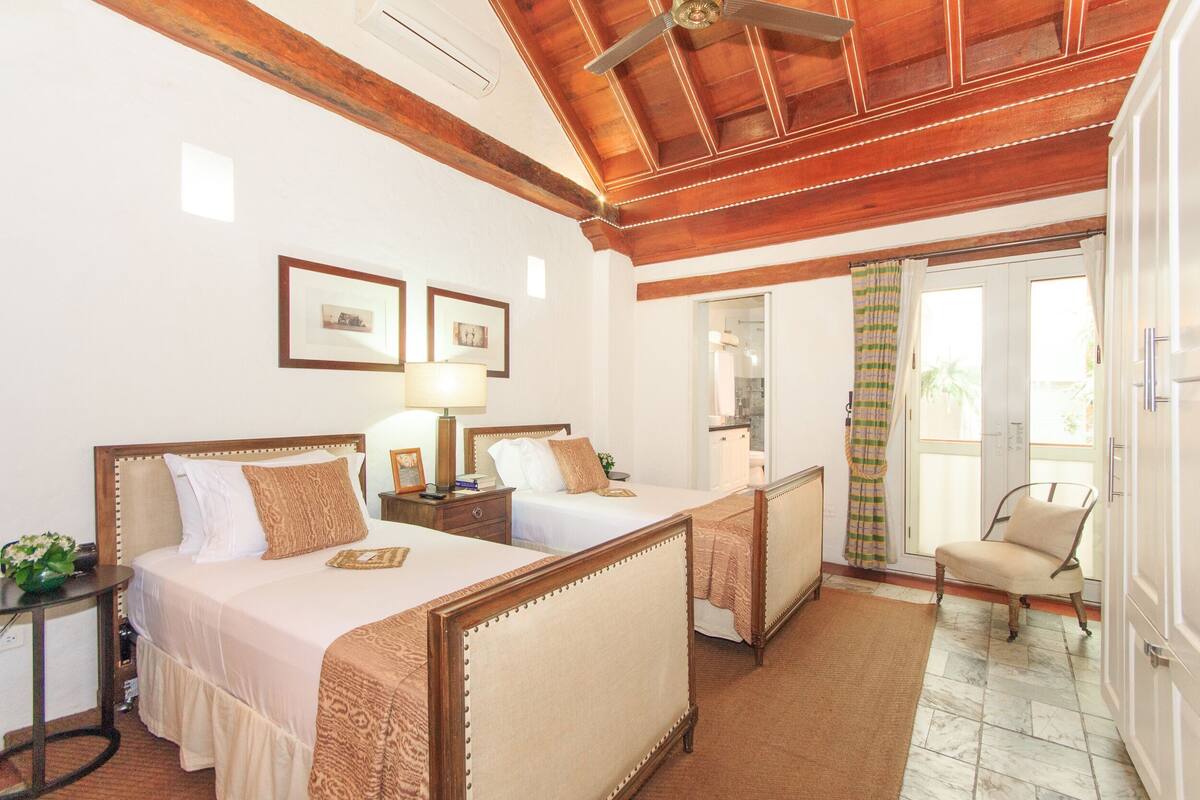 Habitación de hotel con dos camas, ventilador de techo, una mesita con lámpara y una silla.
